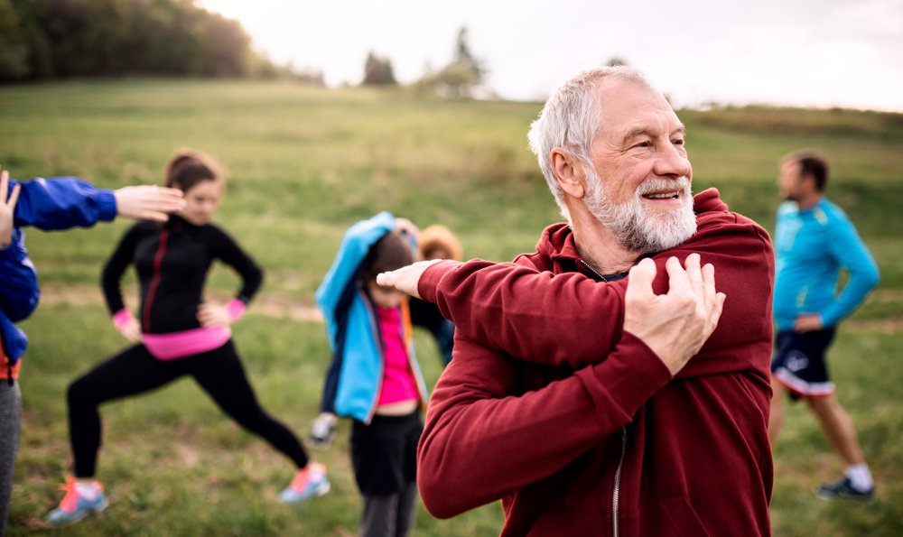 older man stretching outdoors, with other people stretching in the background.