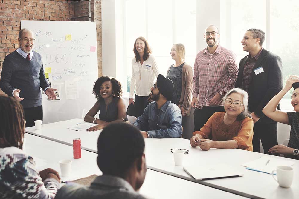 people sitting around a table in a meeting room discussing information on a whiteboard. They are all smiling.