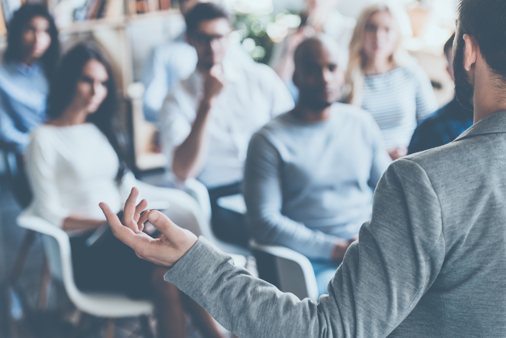 Image shows a man giving a presentation to people in an office