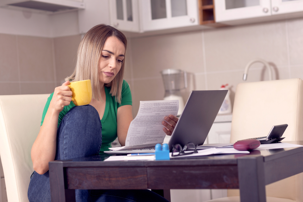 woman assessing her bills, she's holding a cup of coffee and sat on her sofa with her laptop open