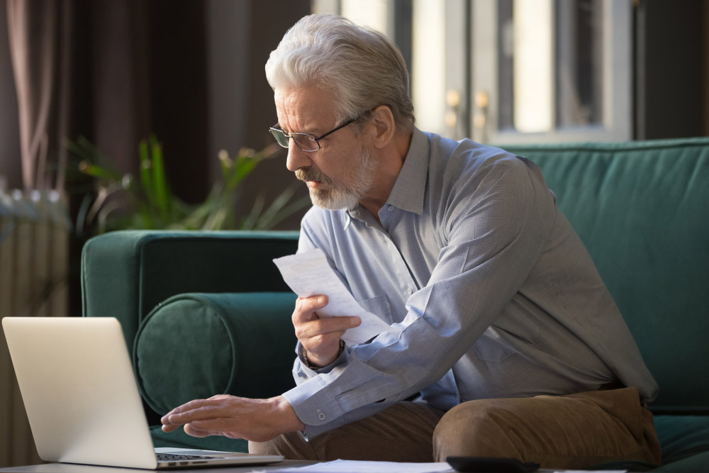 Serious old retired man holding paper calculating paying bills online using looking at laptop, focused senior grandfather make loan bank payment in app internet service on computer sit on sofa alone