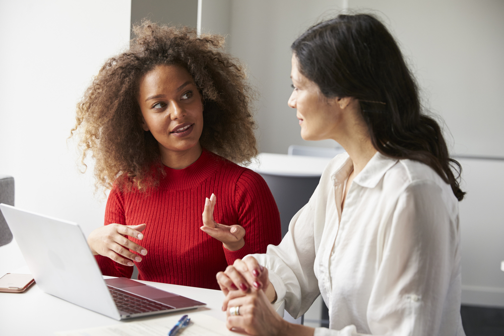 A young black woman is presenting information to another woman at work. They are sat in front of a laptop.