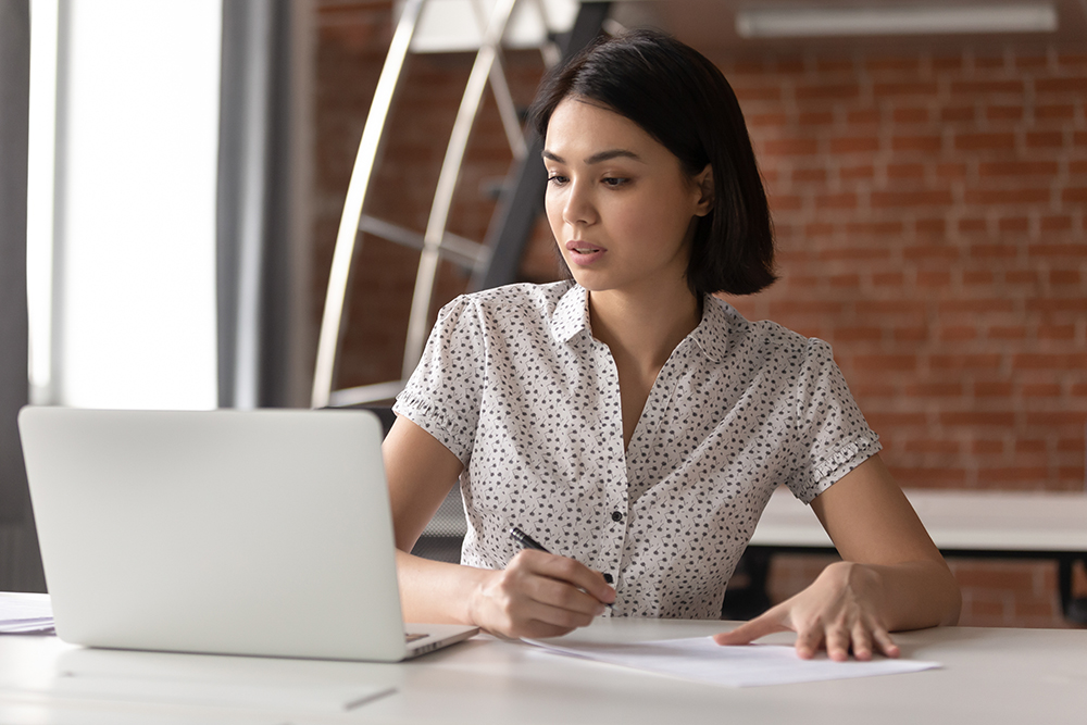 Focused asian business woman working studying online in office looking at laptop making notes, serious japanese employee or student watching webinar writing information in notebook sitting at desk